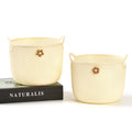 Two cream-colored woven baskets with floral accents on a white background, one on top of a black book titled 'NATURALIS'.